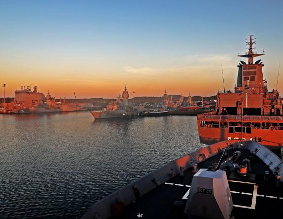 HMAS Choules, HMAS Stuart, HMAS Arunta, HMAS Warramunga, HMAS Toowoomba, HMAS Stalwart and the forecastle of HMAS Hobart in the foreground alongside Fleet Base West as the sun rises.